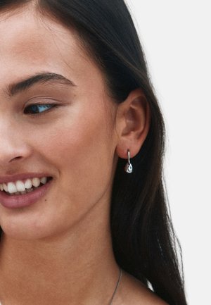 Silverörhängen i hoop-modell med tårformade hängande detaljer som reflekterar ljus, båda bärs av en person med långt mörkt hår och naturlig makeup.