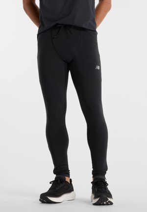 Homme portant des leggings de sport noirs et des chaussures de course noires avec des semelles blanches, debout sur un fond clair uni.