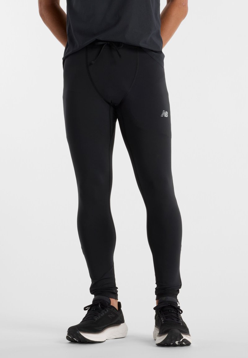 Homme portant des leggings de sport noirs et des chaussures de course noires avec des semelles blanches, debout sur un fond clair uni.