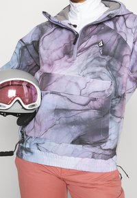 Pull en violet et gris avec un motif, col zippé à mi-hauteur, ourlet élastique et poche avant ; accompagné d'un casque de ski gris avec visière rose.