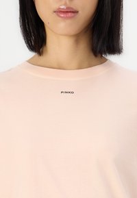Tricou din bumbac roz deschis, cu guler rotund, având un logo negru „PINKO” centrat sub guler. Textură fină, mânecă scurtă.