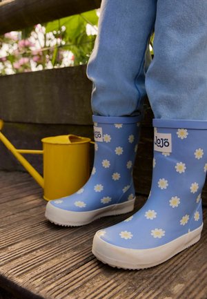 Dítě obuté do modrých gumových bot s bílými kopretinami stojí na dřevěné terase poblíž žluté konev a záhonku.
