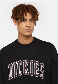 Felpa nera realizzata in morbido tessuto, con collo rotondo e logo "DICKIES" ricamato in bianco e viola scuro.