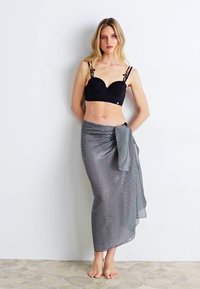 Μαύρο σουτιέν με διακοσμητικές τιράντες, συνδυασμένο με μια μακριά καρό φούστα σε μαύρο και άσπρο, δεμένη κομψά στη μέση. Μοντέλο χωρίς παπούτσια.