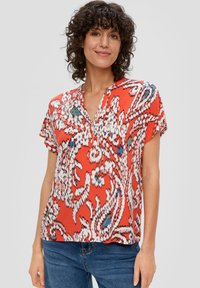 Rote Bluse mit kurzen Ärmeln und V-Ausschnitt, die ein weiß-blaues, abstraktes Paisleymuster aufweist und aus einem weichen, strukturierten Stoff besteht.