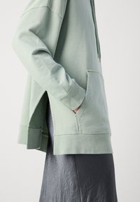 Personne portant un sweat à capuche vert clair avec poche avant et fentes sur les côtés, associé à une jupe texturée foncée.