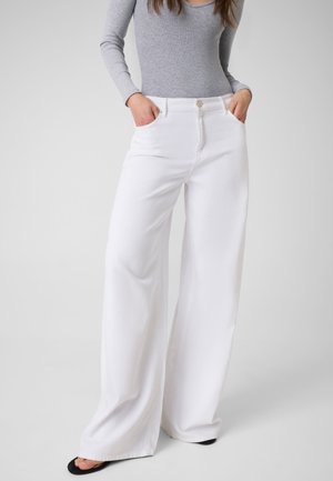 Femme portant un pantalon blanc taille haute à jambes larges avec poches avant, associé à un haut gris cintré à manches longues et des chaussures noires ouvertes à l’avant.
