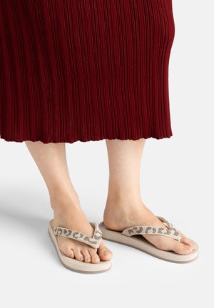 Füße in beigen Flip-Flops mit gemusterten Riemen, gezeigt unter einem roten, gerippten Rock, der bis zur Mitte der Waden reicht, stehend auf einer schlichten Oberfläche.