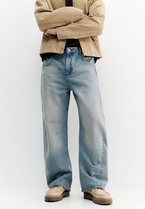 Osoba v béžovej bunde, čiernom tričku, vo voľných svetlomodrých džínsoch a béžových topánkach stojaca so založenými rukami na pozadí bielej steny.