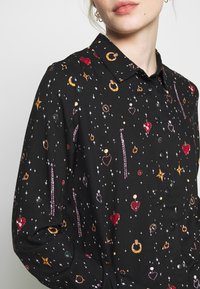Kvinde iført sort skjorte med knapper og et himmel-tema print, der inkluderer stjerner, måner, hjerter, ringe og små ædelsten i forskellige farver.