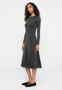 Hosszú ujjú fekete ruha, fodros szegéllyel, csillogó anyagból készült. Texturált felület, kettős ívű élekkel az ujjaknál és a szegélynél.