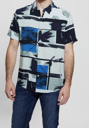 Mann trägt ein kurzärmeliges Hemd mit Knopfleiste und blau-schwarz-grauen abstrakten Blockmustern, kombiniert mit dunkelblauer Jeans.