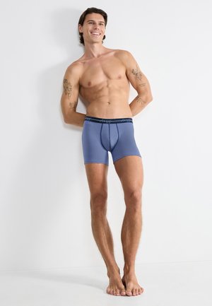 Blue boxer briefs with a black waistband. Smooth texture and fitted design. Displayed on a male model standing against a neutral background.