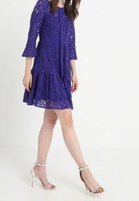 Fialové krajkové šaty s tříčtvrtečními rukávy, vypasovaným pasem, rozšířenou sukní a mašlí v pase; doplněné stříbrnými sandály na podpatku.