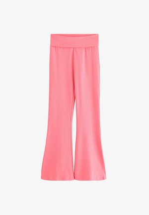 Smilende pige står iført en hvid tanktop, lyserød cardigan, lyserøde bukser og hvide sneakers mod en ensfarvet baggrund.