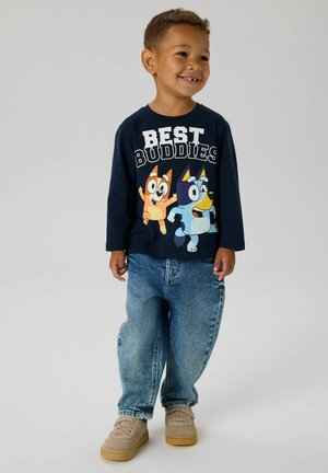 Maglietta a maniche lunghe blu marina con il testo "BEST BUDDIES" e personaggi dei cartoni animati; indossata con jeans baggy azzurri chiari e sneakers beige.