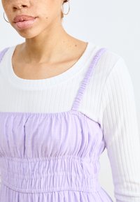 Rochie lavandă cu textură adunată, cu bretele ajustabile, purtată peste un top alb cu mânecă lungă texturat. Guler rotund, material moale.