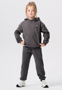 Grijze hoodie en bijpassende sweatpants, gemaakt van zachte stof. De hoodie heeft een voorkantzak en een klein logo. Model draagt witte sneakers.