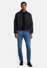 Giubbotto in denim nero con zip frontale, polsini elasticizzati e colletto alto, abbinato a jeans blu e scarpe lace-up nere.