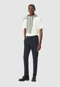 Camicia bianca a maniche corte, maglione grigio drappeggiato intorno al collo, pantaloni blu scuro e scarpe nere. Design semplice e sartoriale con colori solidi.