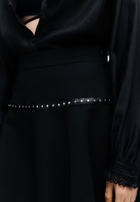Zwarte aansluitende rok met een gladde textuur, voorzien van een glanzende zwarte band die is geaccentueerd met zilveren studs langs de taille. Lange doorschijnende zwarte mouwen met kantenkappen.