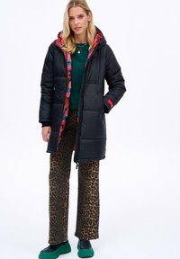 Čierna páperová bunda s kapucňou s kvetinovým vzorom, pod ňou zelený sveter, nohavice s leopardím vzorom a zelené platformové topánky.