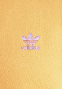 Tejido amarillo con relieve que presenta un logo bordado de Adidas en púrpura claro, con tres hojas y franjas horizontales.