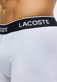 Boxer in cotone azzurro chiaro con fascia elastica nera con la scritta "LACOSTE" in bianco. Texture liscia e design aderente.