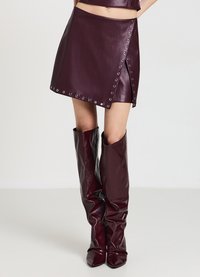 Mini gonna in ecopelle bordeaux con dettagli a grommet, design avvolgente asimmetrico, abbinata a stivali alti fino al ginocchio in tinta unita con punta a punta.
