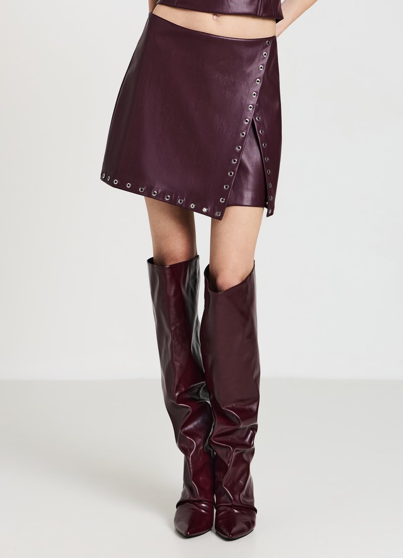 Mini gonna in ecopelle bordeaux con dettagli a grommet, design avvolgente asimmetrico, abbinata a stivali alti fino al ginocchio in tinta unita con punta a punta.