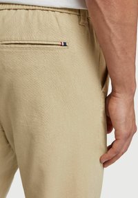 Beige katoenen blend broek met een ontspannen pasvorm, voorzien van een achterzak met een rood, wit en blauw streepdetail. De textuur lijkt glad.