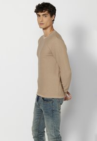 Beige bordázott hosszú ujjú póló, kerek nyakkivágással, testhez simuló fazonnal, világoskék farmernadrággal párosítva.