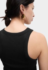 Zwarte geribde tanktop met een brede halslijn en blootgestelde schouderbanden. Gouden statementoorbellen met een gebogen vorm. Glad huidtextuur zichtbaar.