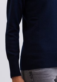 Námorně modrý vlněný svetr s dlouhými rukávy, žebrovanými manžetami a spodním lemem. Texturovaný povrch s přiléhavým střihem, zobrazený vedle šedých džín.