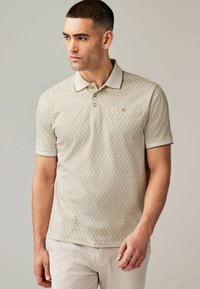 Beige polo-shirt med et geometrisk mønster, korte ærmer og en kontrasterende krave. To knapper ved halsen. Glat stofstruktur.