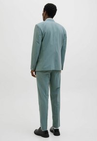 Costum verde deschis cu un design slim-fit, țesătură texturată, rever în formă de cută și închidere cu două butoane. Asortat cu pantofi negri.