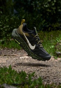 Μαύρο και ελιά αθλητικό παπούτσι με λευκό σήμα, πράσινη σόλα με στίγματα, υφασμένο πάνω μέρος και μαύρα κορδόνια, τοποθετημένο σε φυσικό φόντο.