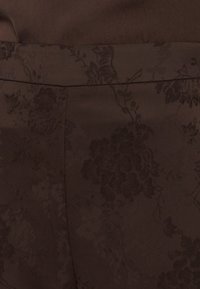 Σκούρο καφέ ύφασμα με διακριτικό φλοράλ σχέδιο ζακάρ και ορατές ραφές, που δείχνουν υφή και λεπτομέρειες σχεδίασης.