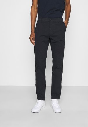 Person, die schwarze gerade geschnittene Hose, weiße Sneakers und ein dunkles Kurzarmshirt trägt, steht vor einem einfachen hellen Hintergrund.