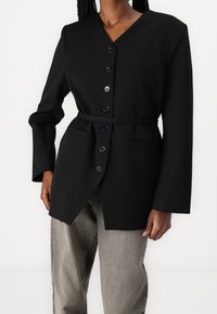 Černý blazer s výstřihem do V, dlouhými rukávy, šesti předními knoflíky a opaskem s mašlí v pase. Obsahuje dvě boční kapsy a má upravený střih.