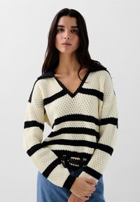Pull en tricot rayé crème et noir avec un col en V, des dentelles et un col. Associé à un jean bleu taille haute et une ceinture à motifs.
