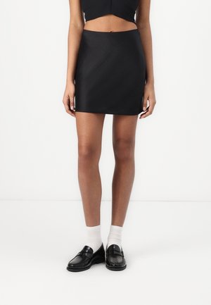 Femme portant une mini-jupe noire, un top court noir, des chaussettes blanches aux chevilles et des mocassins noirs, debout devant un fond blanc uni.