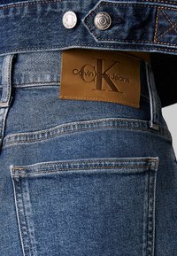 Vaqueros de mezclilla en un lavado azul medio, con una etiqueta de cuero marrón que dice "Calvin Klein Jeans" grabada, y bolsillos traseros cosidos con costuras visibles.