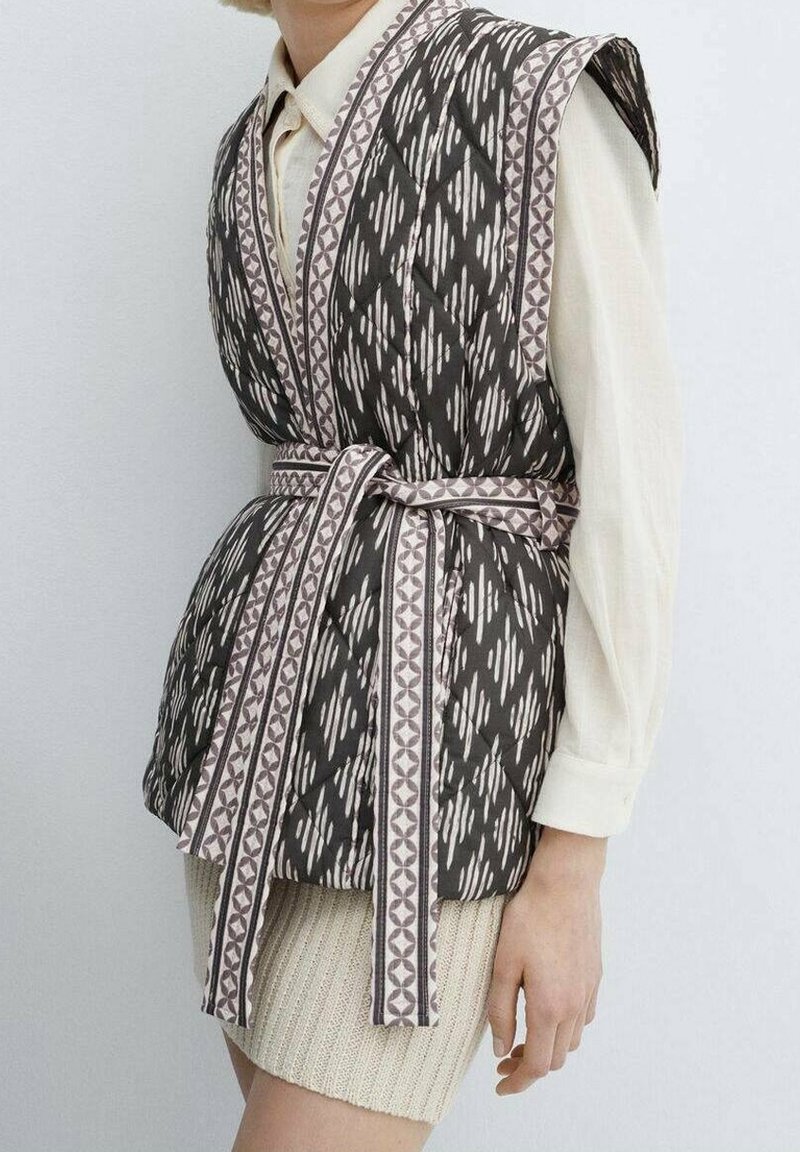 Gilet sans manches à motifs noir et blanc avec ceinture porté sur une chemise crème à manches longues et une jupe en tricot beige sur le torse d'une personne.
