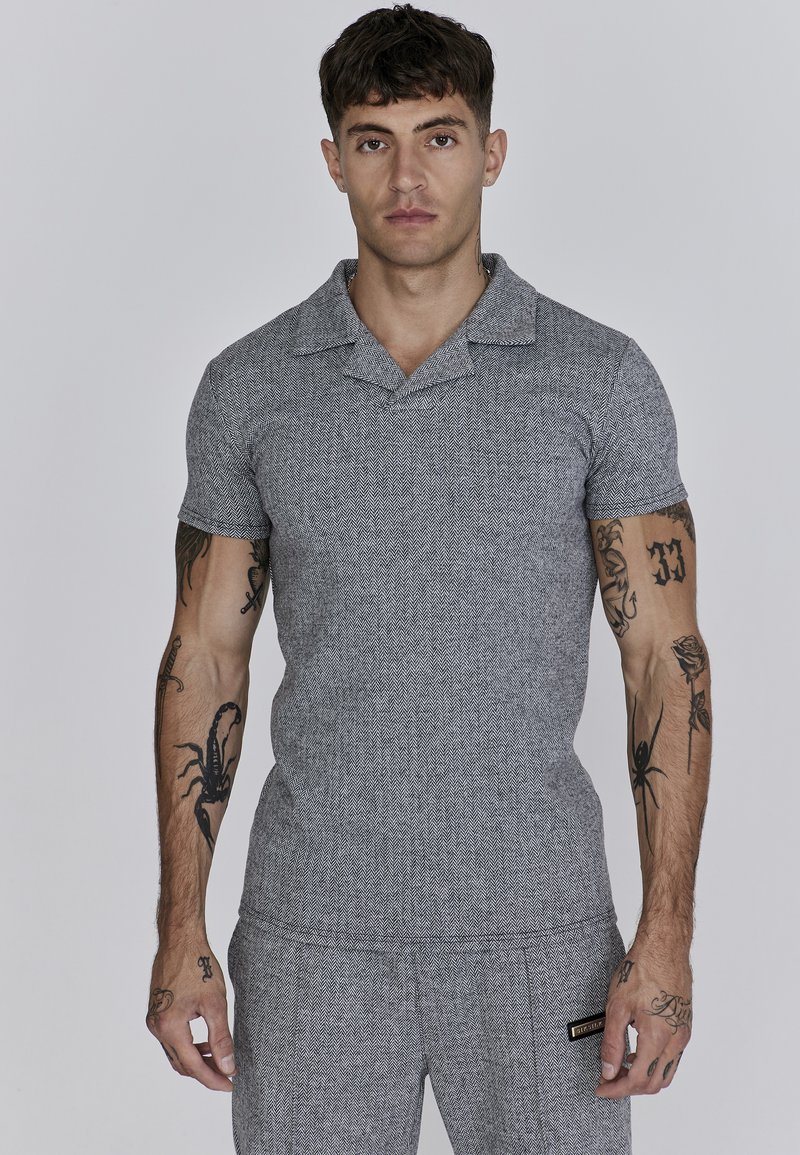 Šedý herringbone polo tričko s šálovým límcem, přiléhavý střih, krátké rukávy a jemnou strukturu. Na boku se nachází malá nášivka značky.