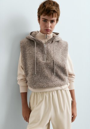 Junge Person mit kurzen Haaren, die eine beige Fleeceweste mit Reißverschluss und Kapuze über einem hellen Langarmshirt und cremefarbenen Hosen trägt.