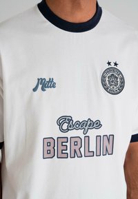 White t-shirt with black collar and sleeves, featuring text "Matte," "Escape BERLIN," and a circular emblem with two stars and the words "Prohibited Berlin."