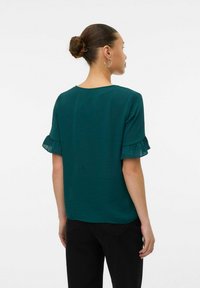 Top corto a maniche corte in teal con maniche trasparenti a volant, vestibilità rilassata e collo rotondo. Tessuto morbido, vista posteriore, abbinato a pantaloni neri.