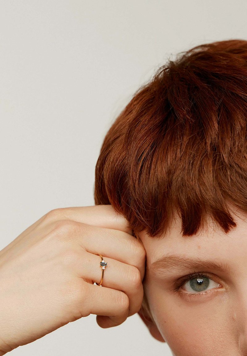 Anillo de oro con una piedra rectangular, llevado en una mano que reposa sobre una cara. Se pueden ver parcialmente unos cabellos cortos y castaños y unos ojos verdes.