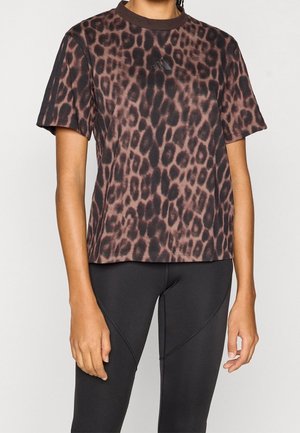Personne portant un t-shirt Adidas marron et noir à imprimé léopard et un legging de sport noir, debout devant un fond clair.